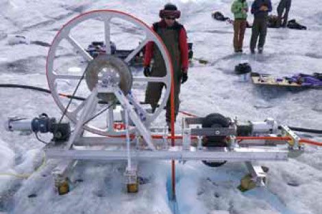 Using a jet of high-pressure, near-boiling water, Harper and his crew drill a borehole through the Greenland ice sheet. The drill lowers a long stem down the borehole until it reaches the ice sheet’s bed.